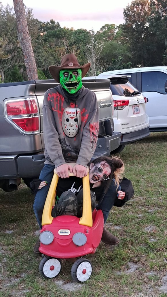 Gideon the Skeleton pushes another actor sat in a children's toy car