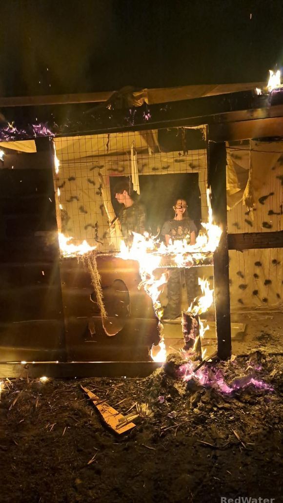 It appears that two people are standing inside a small structure as it burns, they are actually behind the structure visible throug a window